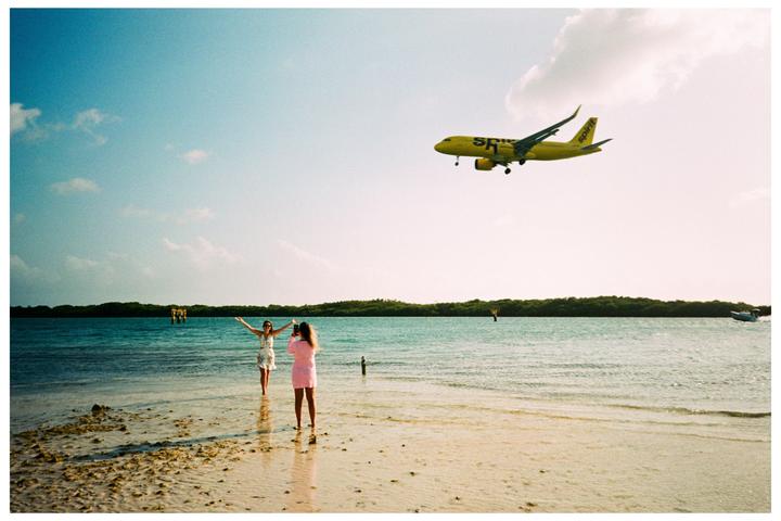Two women stand on a beach with a yellow airplane flying overhead.