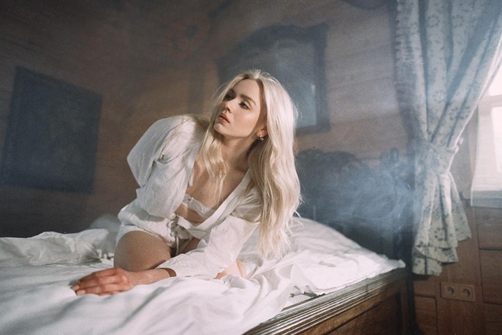 A woman with long blonde hair kneels on a bed in a room with light rays visible in the hazy air.
