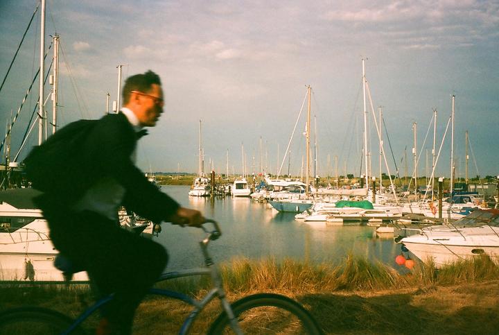 A man in a suit rides a bicycle past a marina filled with boats.