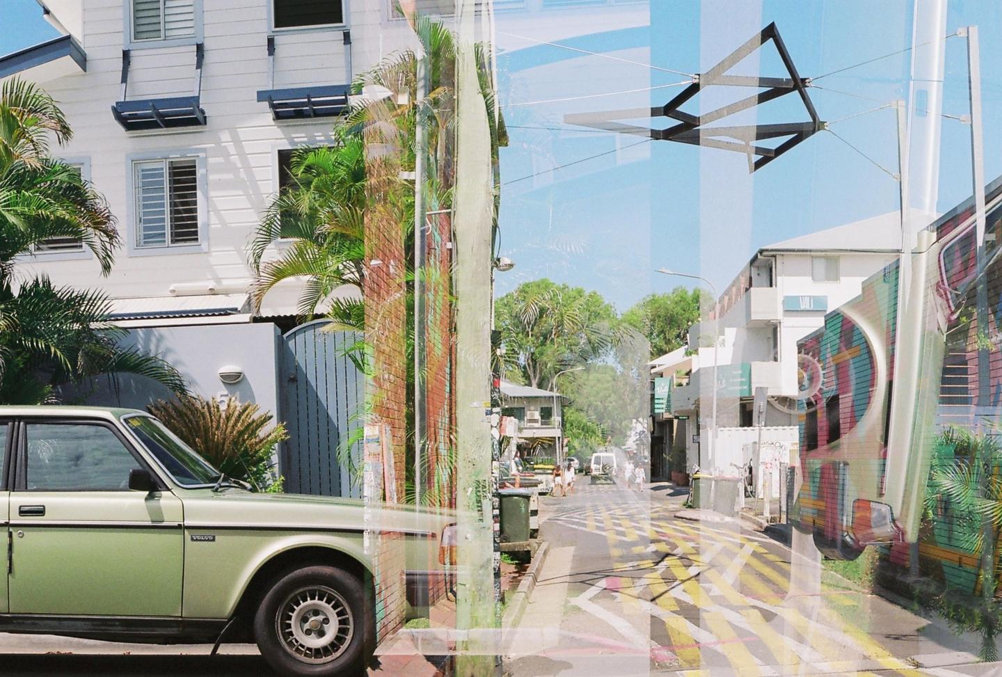 A double exposure shows a green car parked in front of a white building and a street with yellow markings.