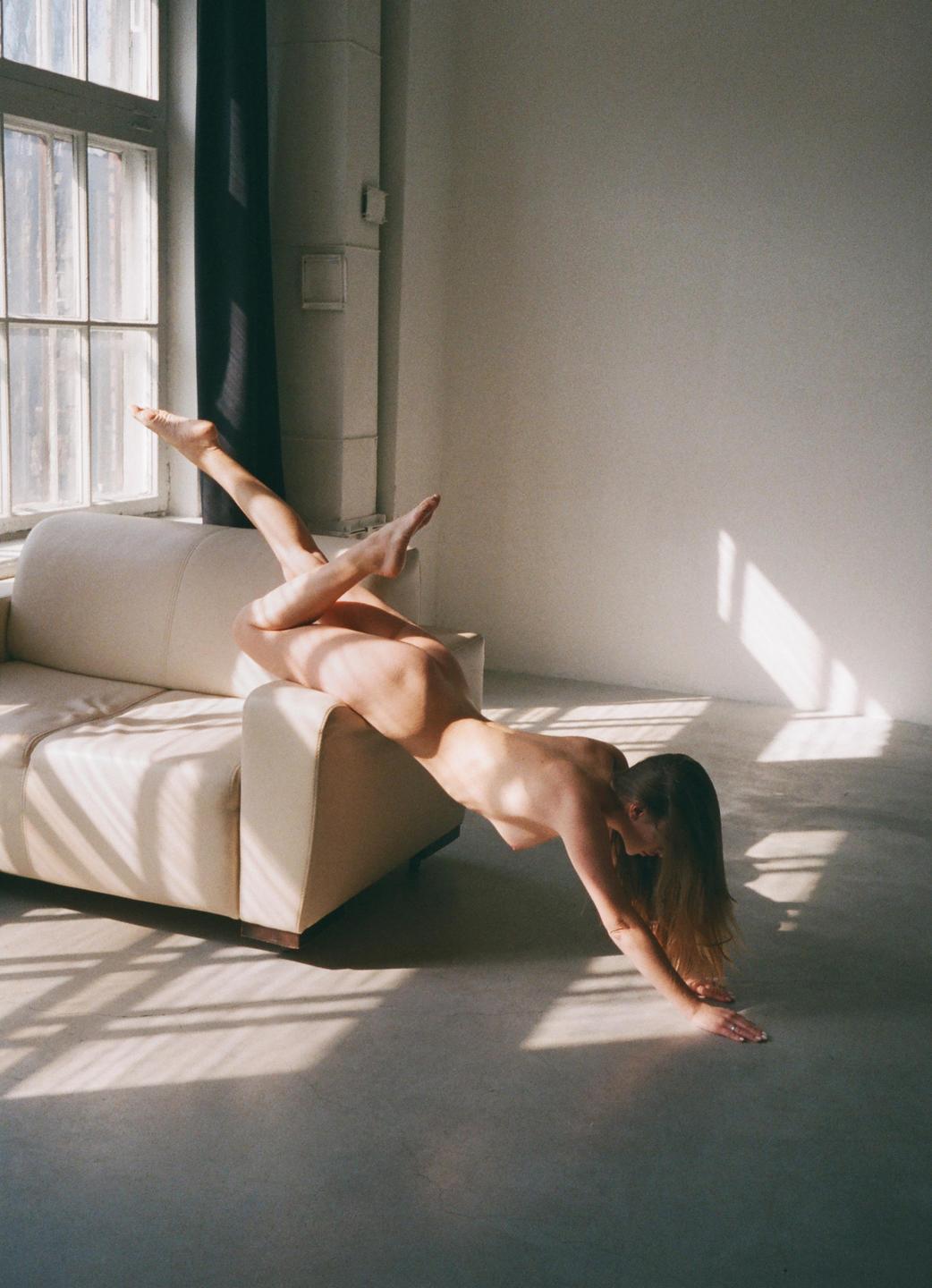 A nude woman is posed on a sofa with her legs in the air and her hands on the floor, with window light creating shadows on the wall and floor.
