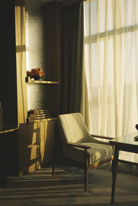 Sunlight streams through a sheer curtain onto a chair and a bookshelf with books and a floral arrangement.