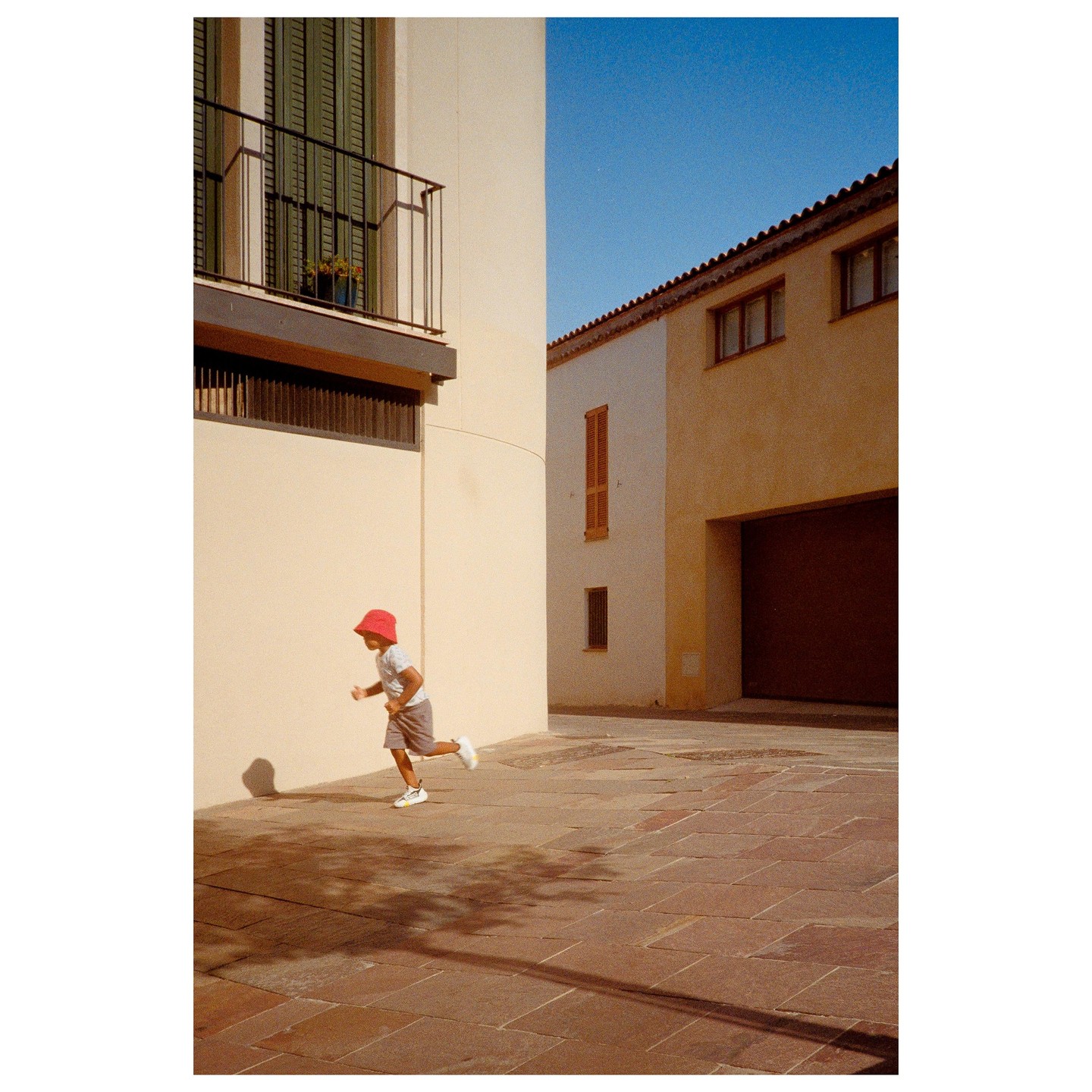 A child in a red hat runs on a paved street between buildings.