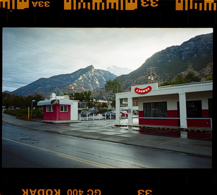 A hot dog shop with a red and white exterior stands beside a wet road, with mountains in the background under a cloudy sky.