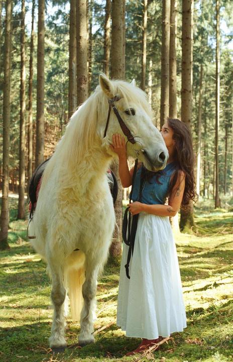 A woman in a white skirt and denim top stands next to a white horse in a forest.