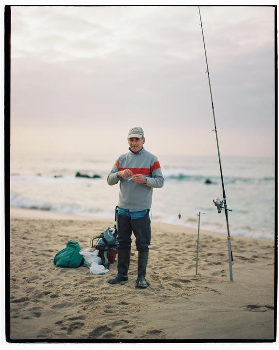 A man stands on a sandy beach holding a small object, with a fishing rod and the ocean in the background.
