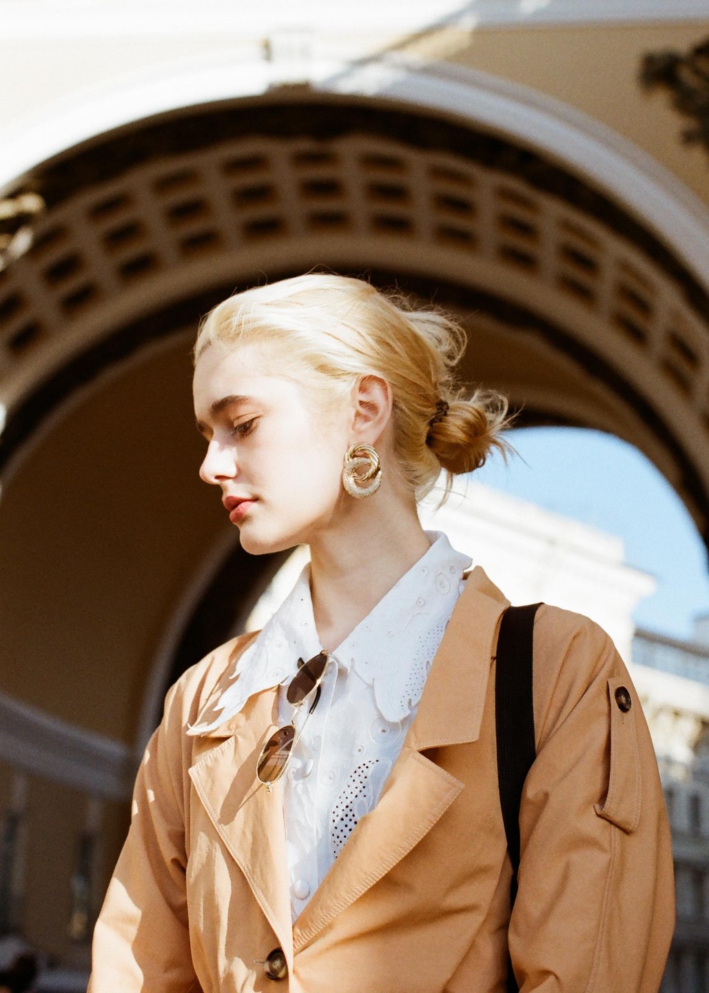 A woman with blonde hair and a tan coat stands in front of an arched building.