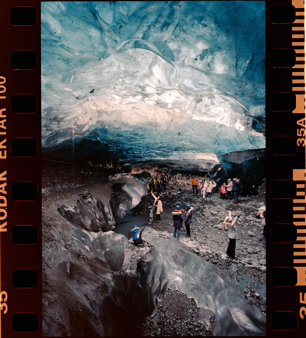 A group of people stand inside a large ice cave with blue and white ice formations.