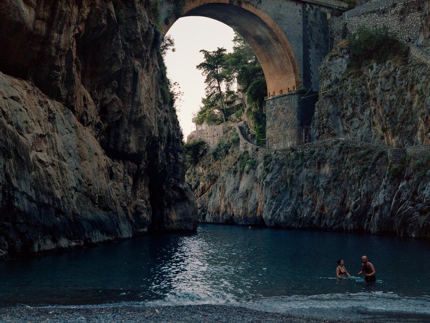 A couple wades in the water under a large stone bridge and rocky cliffs.