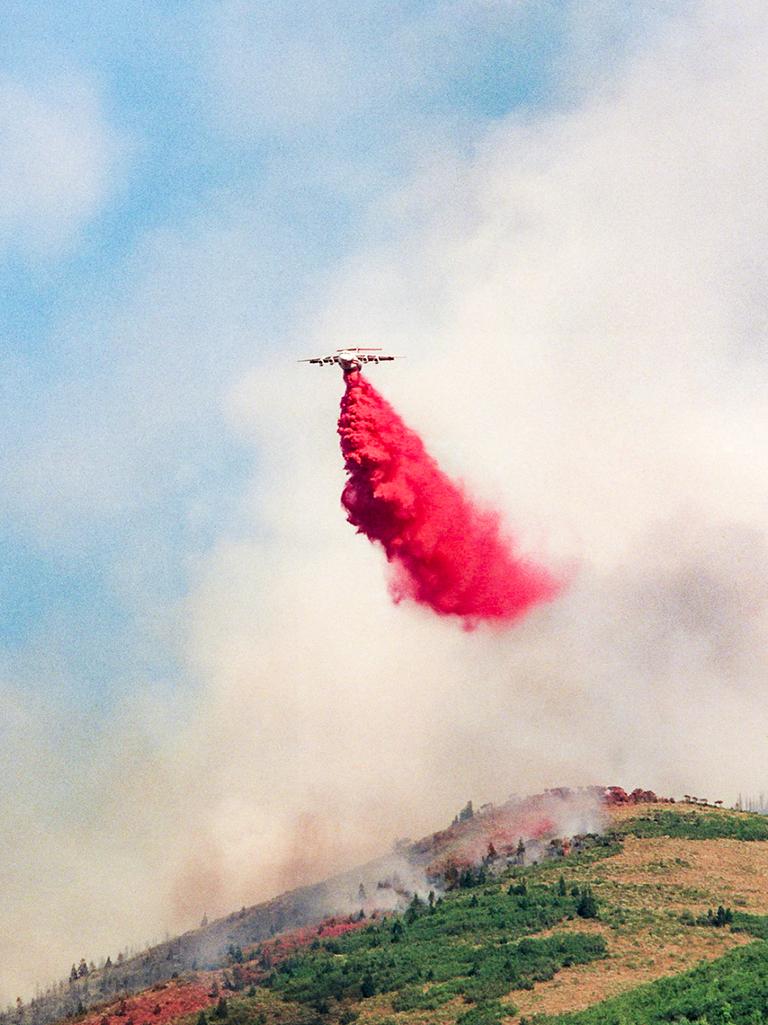 An airtanker drops red fire retardant over a smoky, forested hillside.