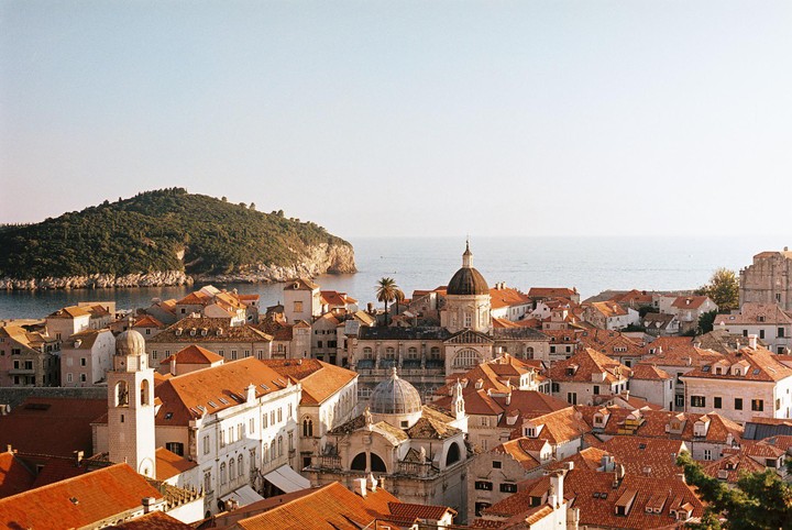 An aerial view shows a city with terracotta roofs and domed buildings next to a body of water and a large, tree-covered island.