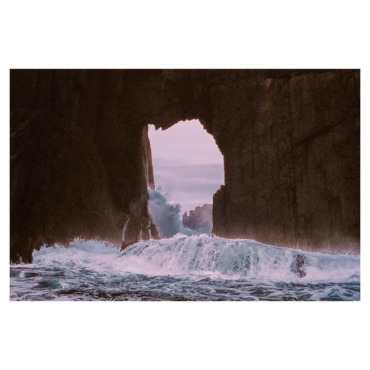 Waves crash through a natural rock archway, revealing a distant coastline under a hazy sky.