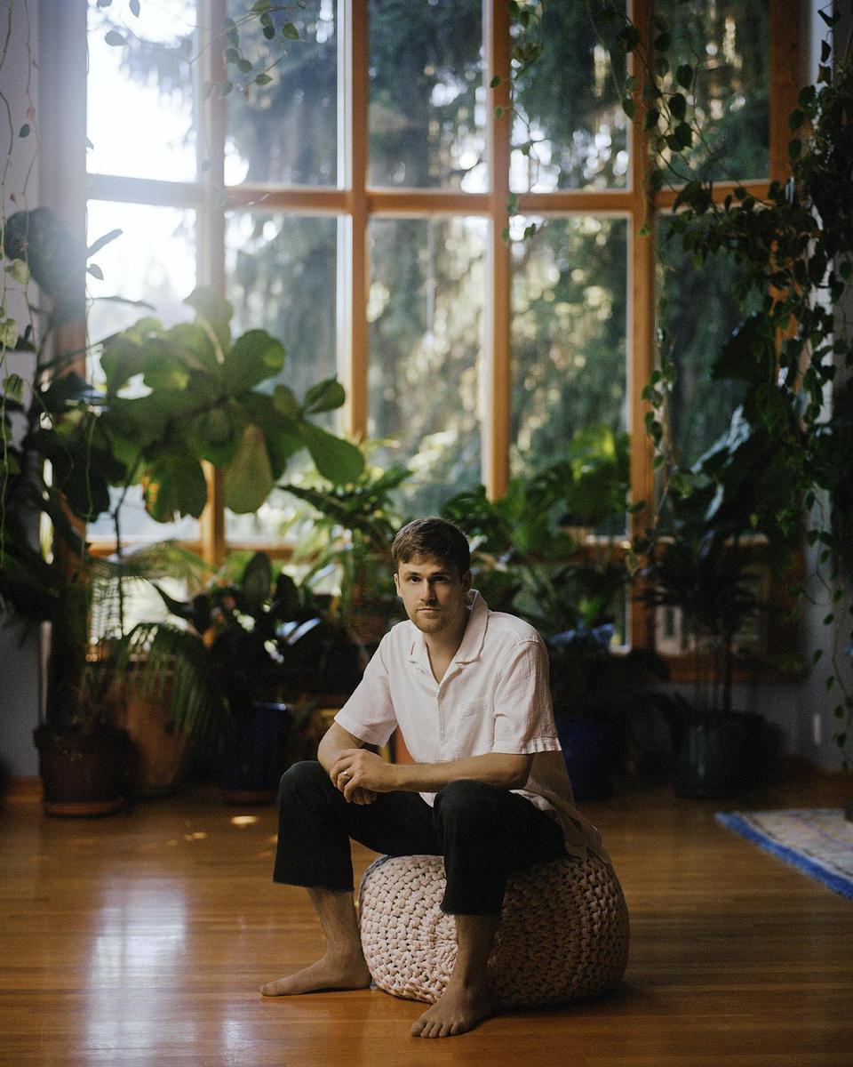 A man sits barefoot on a knitted pouf in a room filled with plants and natural light.