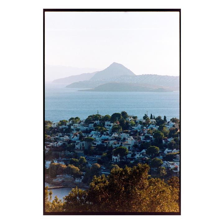 A coastal town with white buildings and green trees sits on a hillside overlooking a body of water with mountains in the distance.