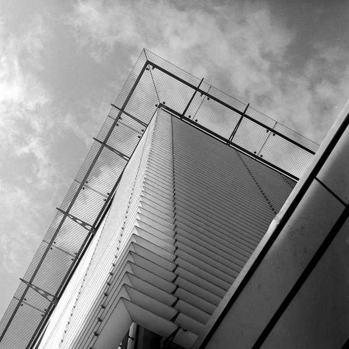 A low-angle view shows the corner of a modern building with a grid-like structure on its roof against a cloudy sky.