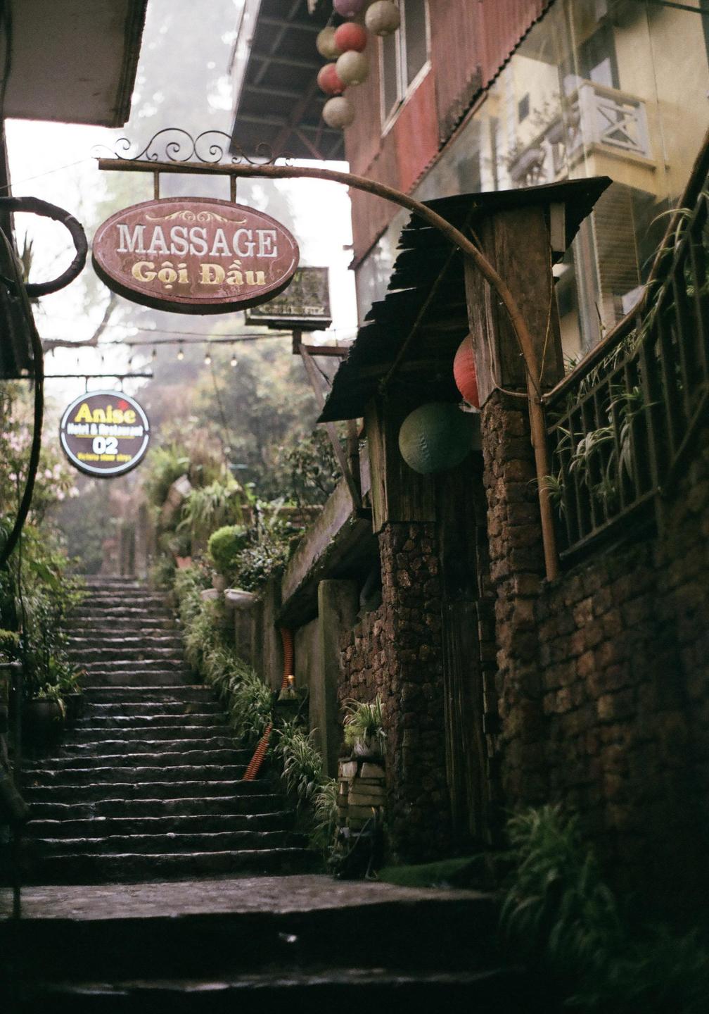 A narrow, steep stone staircase leads up a misty alleyway lined with buildings and lush greenery.