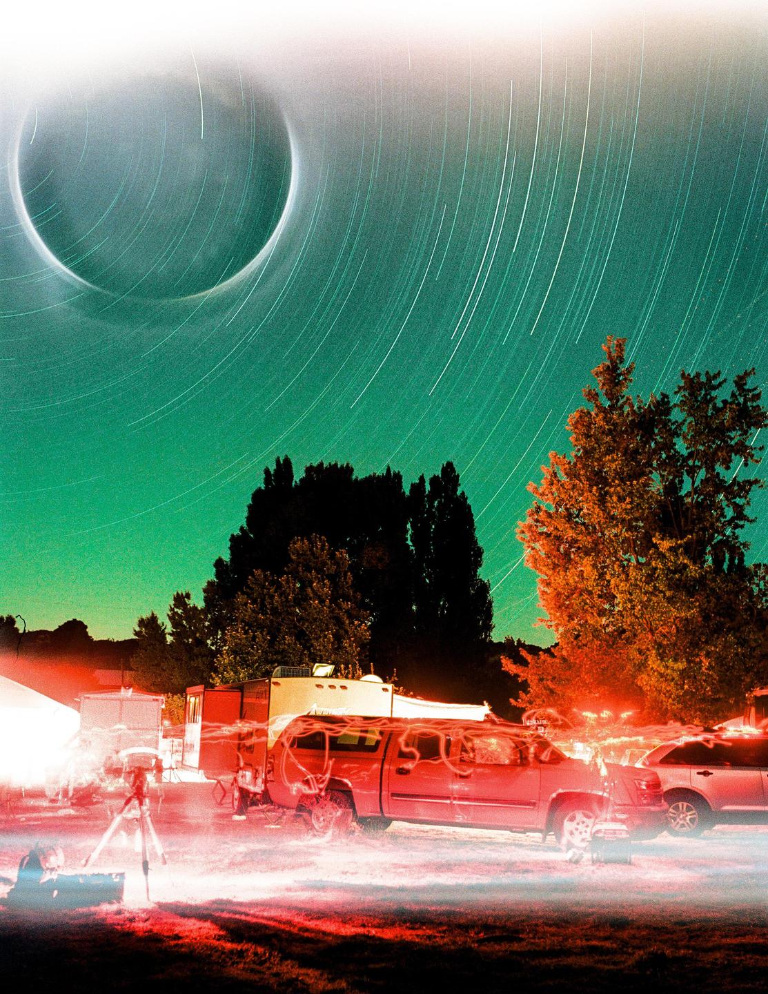 A long exposure photograph shows star trails and a solar eclipse in the sky above a campsite with vehicles and trees.