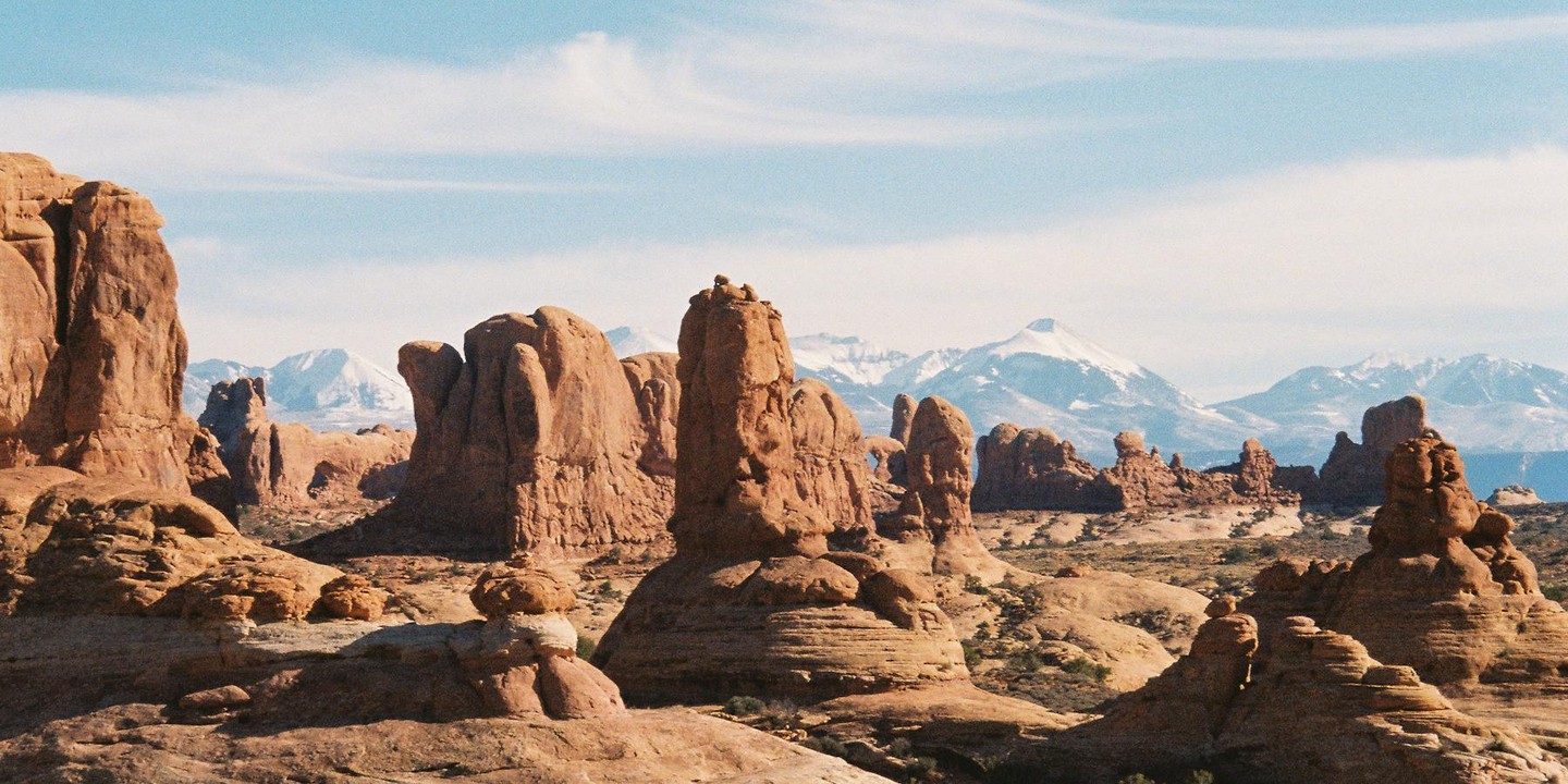 A vast desert landscape features red rock formations under a blue sky with snow-capped mountains in the distance.