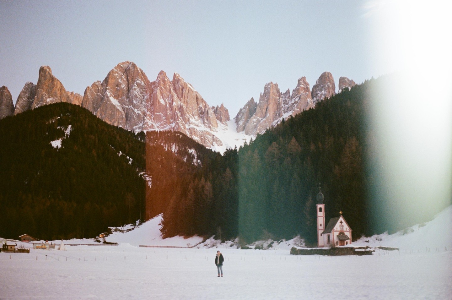A person stands in a snow-covered field with a small church and mountains in the background.