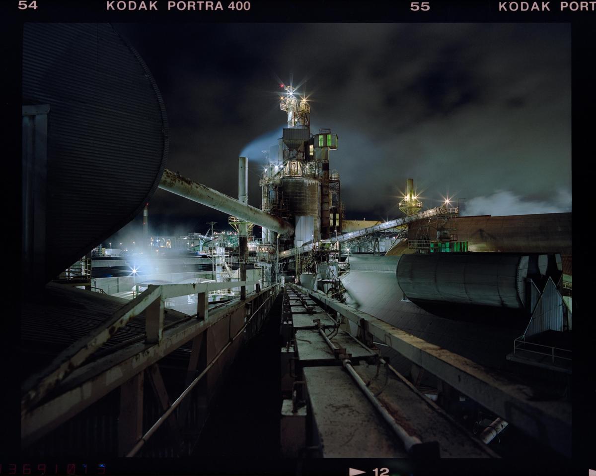 A large industrial plant with multiple structures, pipes, and conveyors is illuminated at night, with some steam or smoke visible.