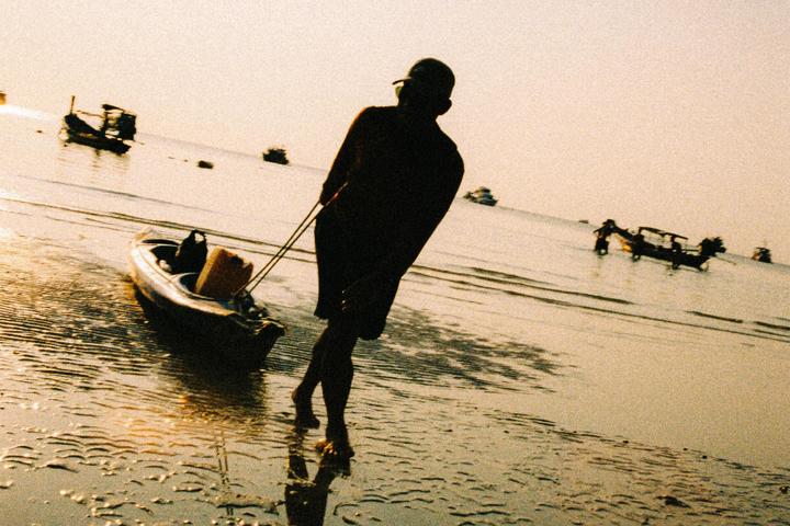 A man pulls a kayak along a sandy beach with boats in the background.