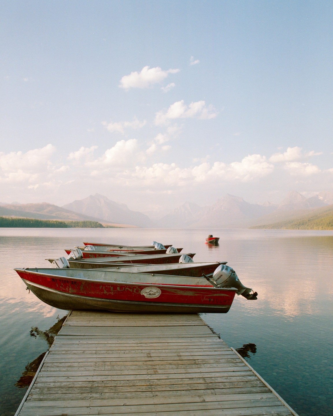 Several red boats are docked at a wooden pier on a lake with mountains in the background.