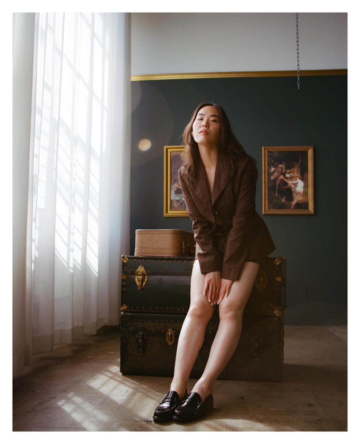 A woman in a brown blazer and loafers sits on stacked trunks in a room with paintings on the wall and light streaming through a window.