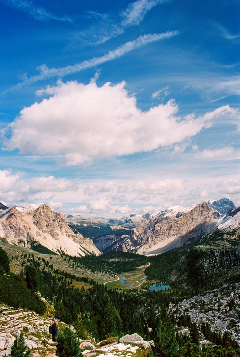 A person stands on a rocky mountain path overlooking a valley with lakes and distant peaks under a blue sky with clouds.