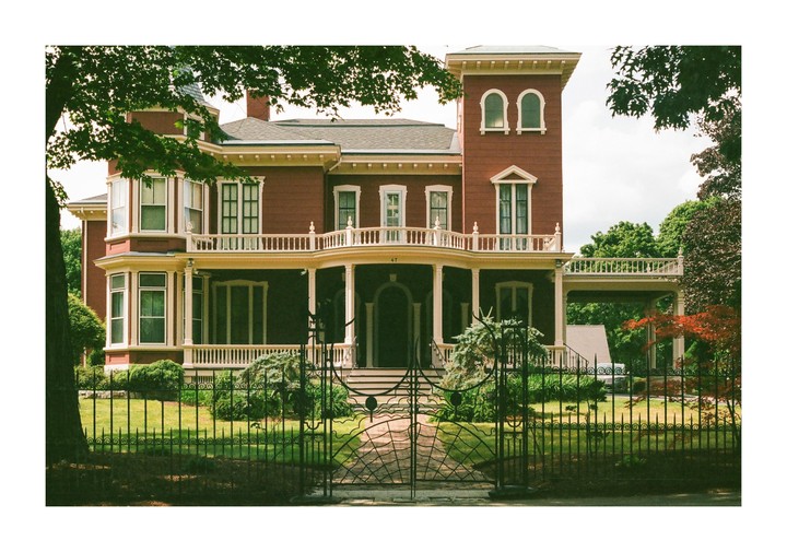 A large red house with a white porch and a black metal fence stands behind a green lawn.