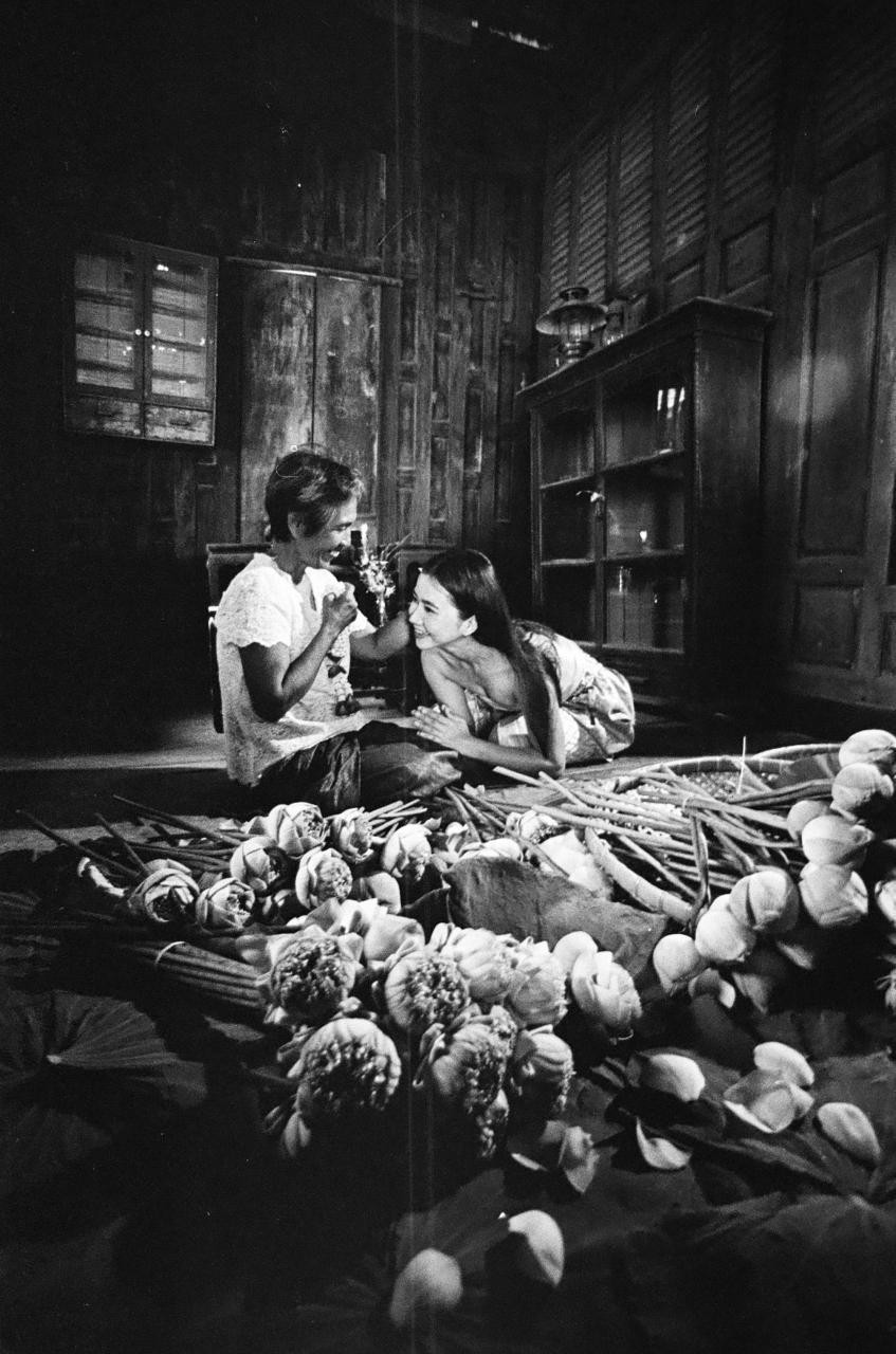 Two women are sitting on the floor surrounded by lotus flowers and stems.