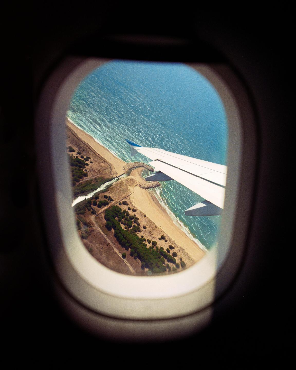 An airplane wing is visible through a window, flying over a coastline with a beach and ocean.