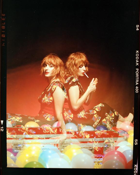 A woman with red hair is sitting back to back with her double exposure, who is lighting a cigarette, surrounded by colorful balloons.
