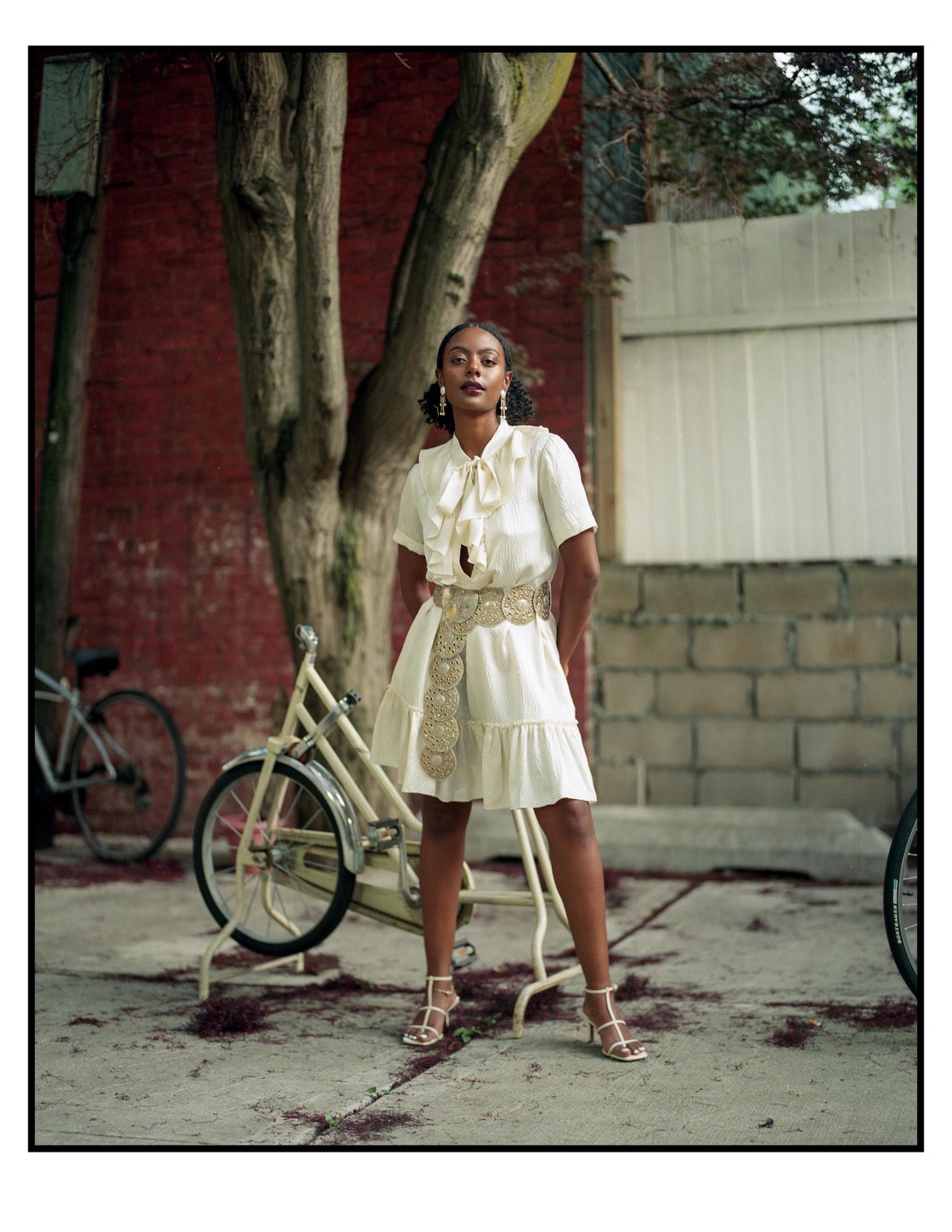 A woman in a white dress and gold belt stands on a paved surface next to a bicycle.