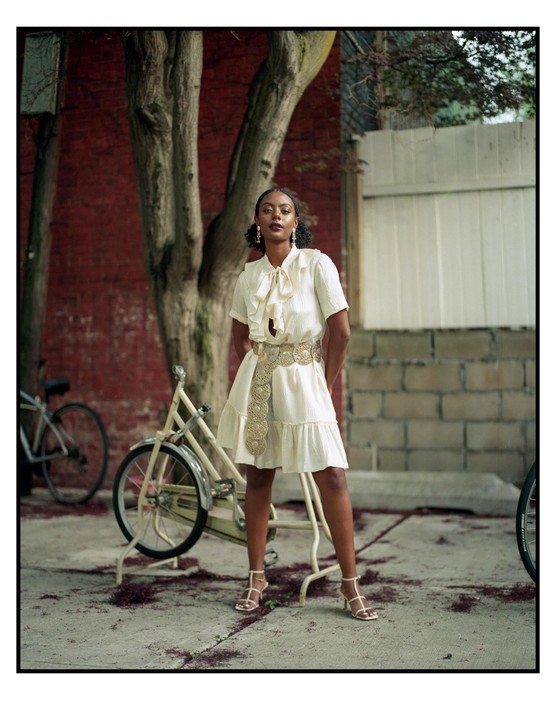 A woman in a white dress and gold belt stands on a paved surface next to a bicycle.