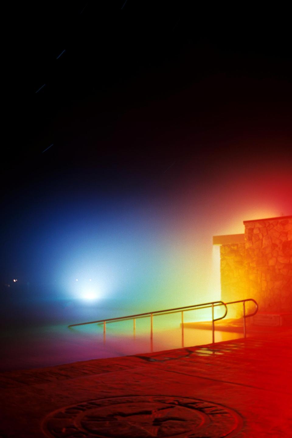 A brightly lit swimming pool with a stone building and railing is visible at night.