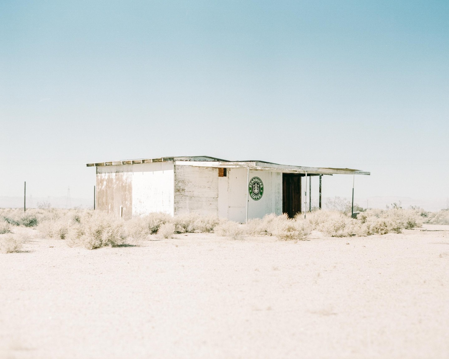 An abandoned white building with a green Starbucks logo on its side stands in a desert landscape under a clear sky.