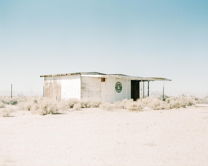 An abandoned white building with a green Starbucks logo on its side stands in a desert landscape under a clear sky.