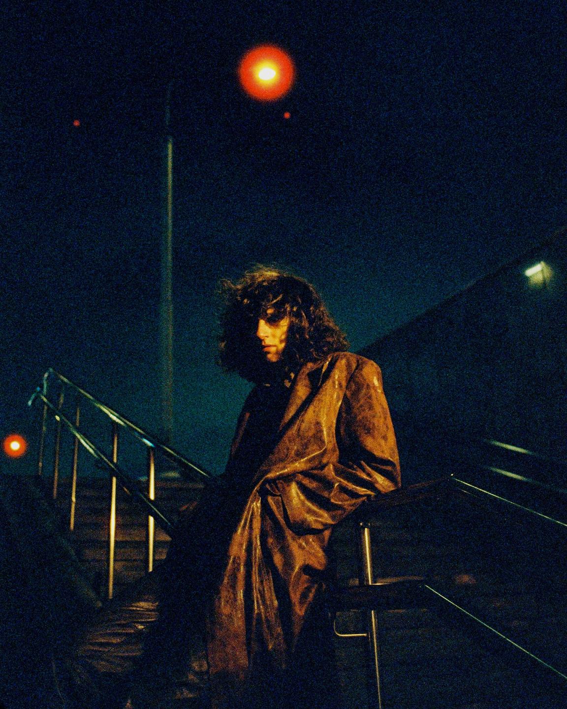 A man with long hair and a brown coat leans on a railing at night.