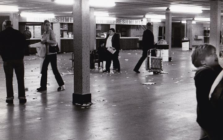 People walk across a dirty floor in a large room with refreshment stands.