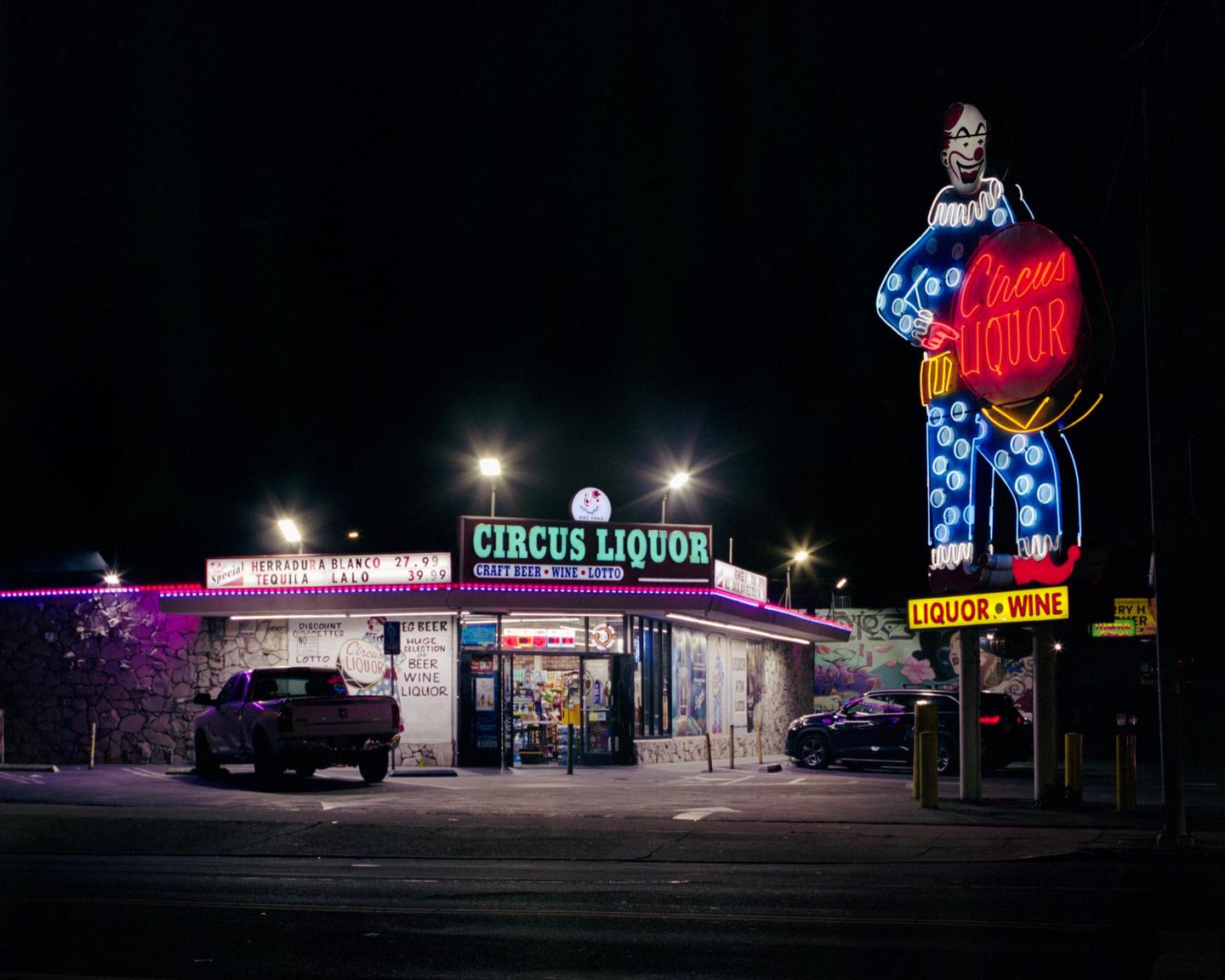A liquor store with a large clown neon sign stands illuminated at night.