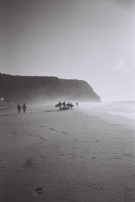 A group of surfers with their boards stands on a beach with a cliff in the background.