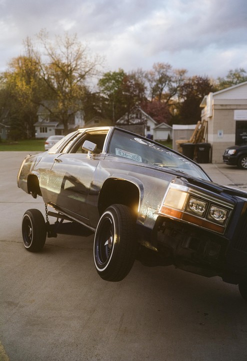 A dark blue lowrider car is shown with its front wheel lifted off the ground.