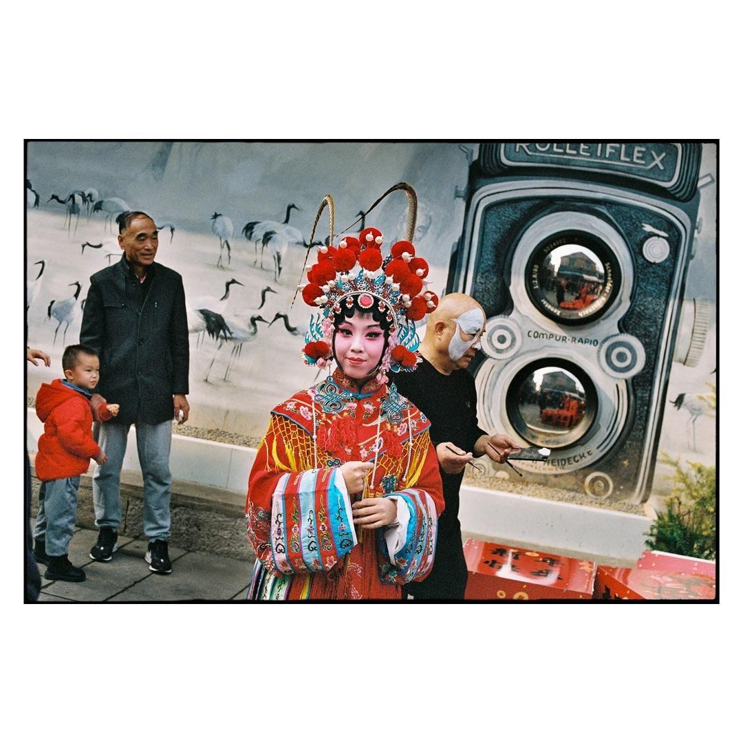 A woman in traditional Chinese opera attire stands in front of a mural of cranes and a large camera, with a man and a boy nearby.