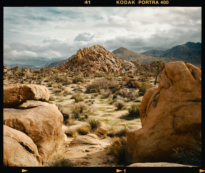 A desert landscape features large rock formations, sparse vegetation, and mountains under a cloudy sky.