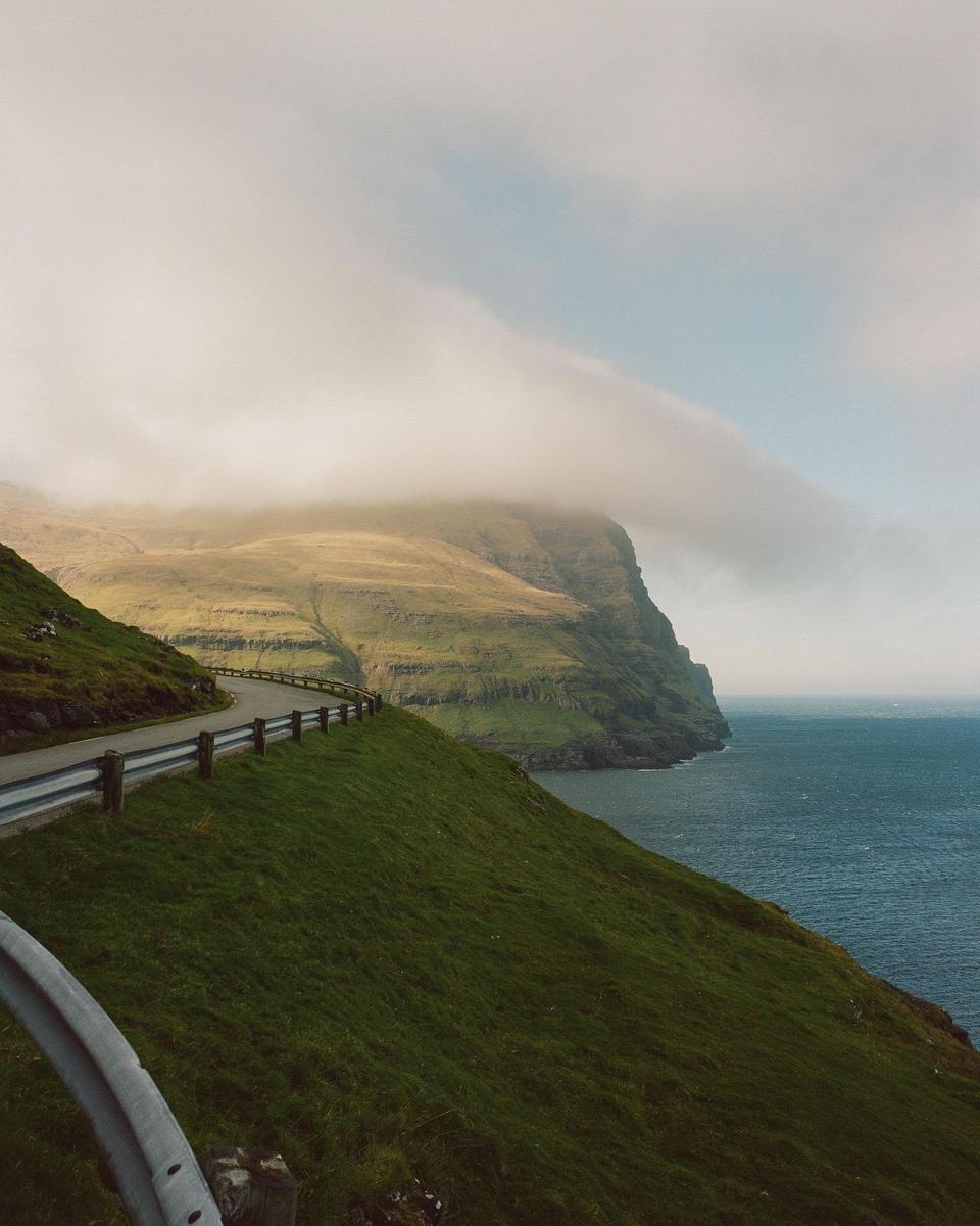 A winding road with a guardrail runs along a grassy hillside next to a body of water, with a large mountain partially obscured by fog in the background.