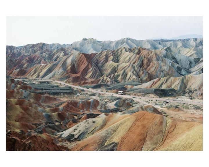 A white van drives along a road through colorful, layered mountains.