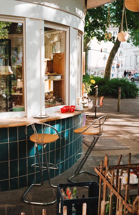 A small outdoor bar with two stools sits on a tiled patio next to a street with trees and buildings.