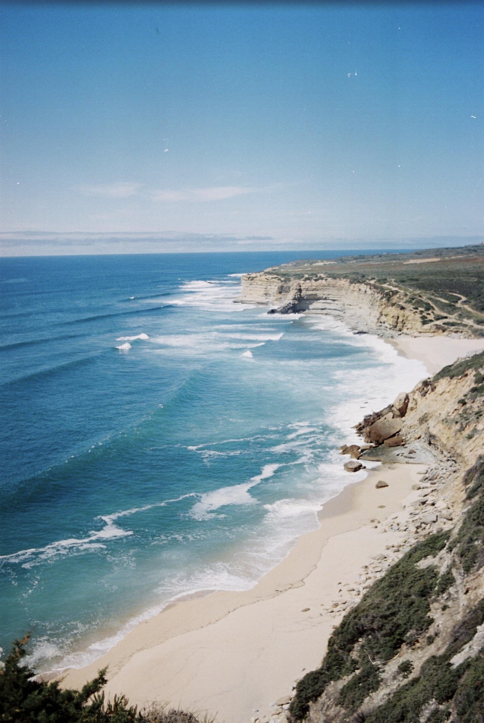 A long stretch of sandy beach meets the ocean with waves breaking on the shore, backed by a tall cliff.