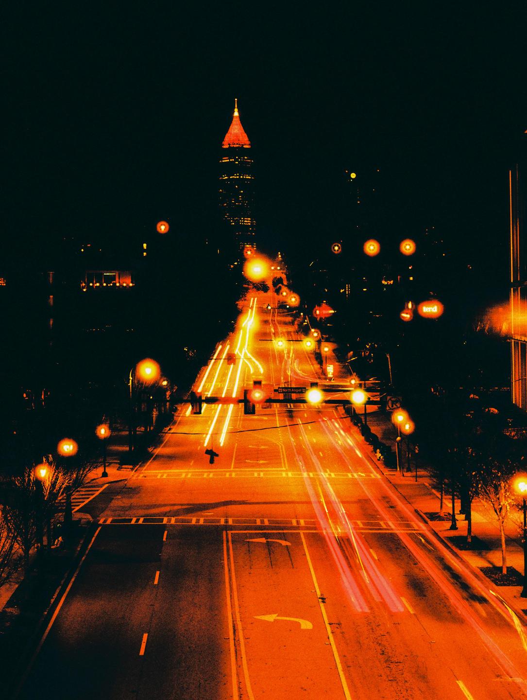 A long exposure shot of a city street at night shows car light trails leading towards a tall building with a pointed, illuminated red roof.