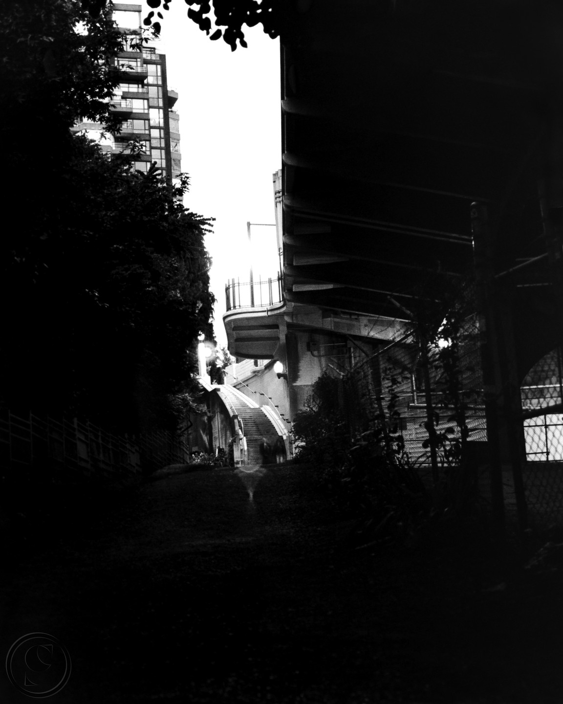 A dark, low-angle view shows a path leading to a brightly lit staircase between a tall building and an overpass.